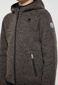 Grå texturerad jacka med huva, som har en framtida dragkedja, sidofickor och ett märke på vänster ärm.