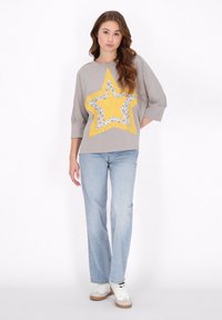 Grå pullover med et stort gult stjernedesign, der har blomstermotiver. Sammen med lyseblå jeans og hvide sneakers.