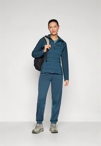 Τζάκετ σε αποχρώσεις τιρκουάζ με φερμουάρ και επένδυση στο μπροστινό μέρος, συνδυασμένο με παντελόνι στο ίδιο χρώμα. Το μοντέλο κρατάει μαύρο σακίδιο πλάτης και φοράει γκρι αθλητικά παπούτσια. Ομαλή υφή.