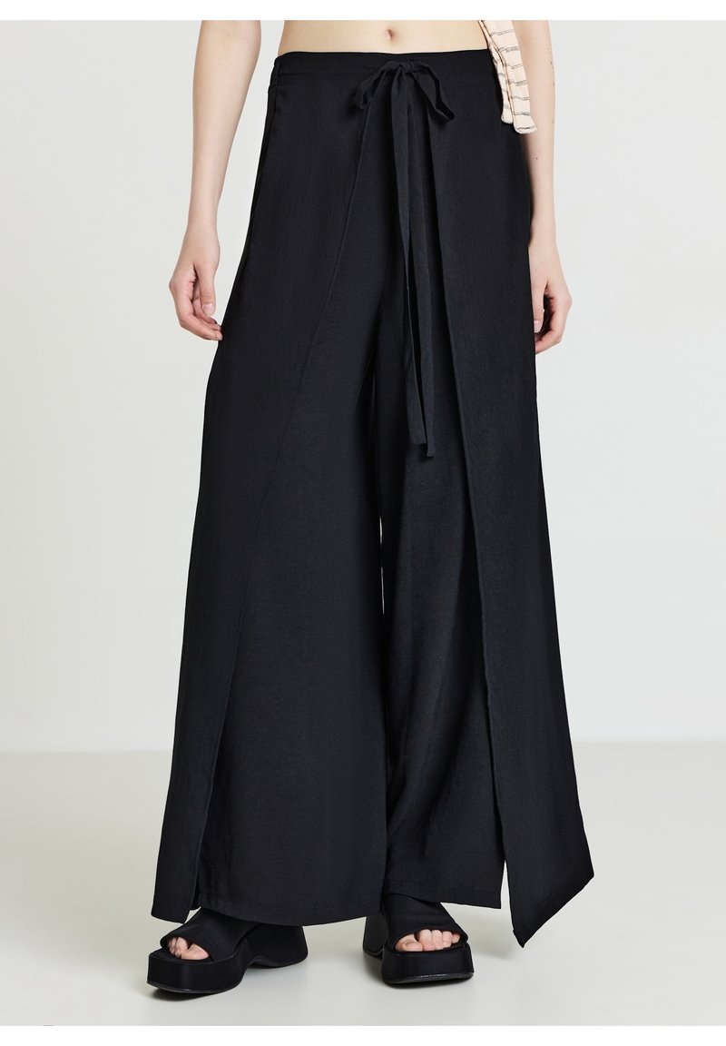Pantalon noir à jambes larges avec détail noué devant, porté avec des sandales à plateformes noires et un top court visible de la taille aux pieds.