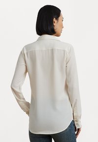 Cream-colored, long-sleeve shirt with a collar, buttoned cuffs, and a curved hem. Made of lightweight fabric, featuring a smooth texture.