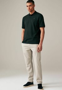 Polo shirt verde oscuro de manga corta y con cuello, combinado con pantalones beige claros y zapatillas negras. Logotipo dorado notable en la camiseta.