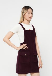 Robe salopette en velours côtelé bourgogne avec deux poches avant, bretelles réglables et une silhouette ajustée. Portée par-dessus un T-shirt blanc.