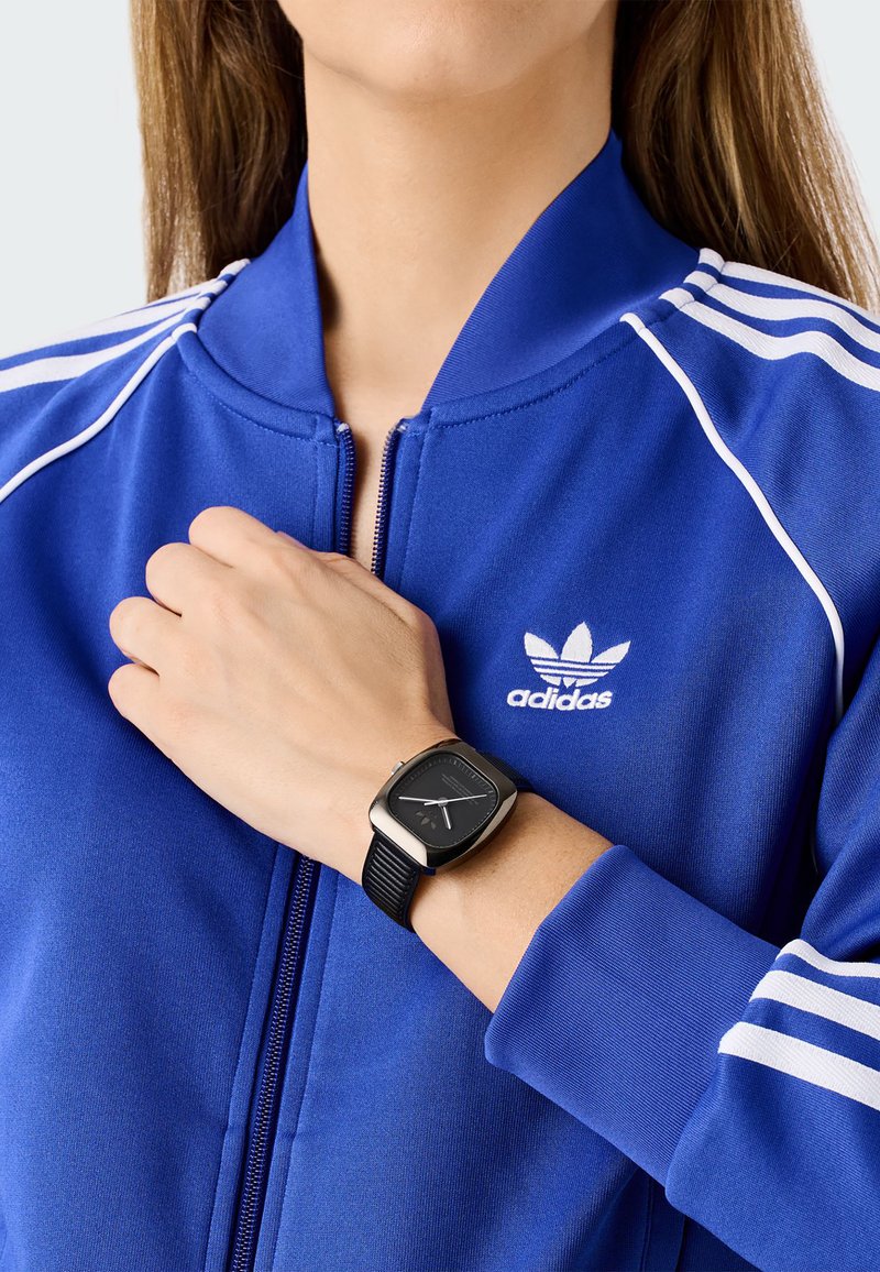 Reloj cuadrado negro con una correa texturizada, llevado en una muñeca, combinado con una chaqueta azul con cremallera que presenta rayas blancas y un logo.