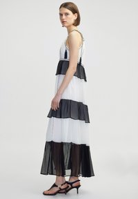 Maxikleid aus weißem und schwarzem Stoff, mit gestuften Lagen, einer definierten Taille und Quastenakzenten, ergänzt durch schwarze Sandalen mit Riemchen.