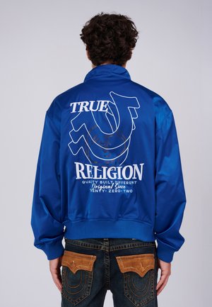 Man met een koningsblauwe jas met witte "True Religion" tekst en logo op de rug, gecombineerd met donkere spijkerbroeken met bruine zakdetails.