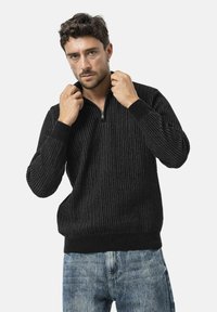 Zwarte geribde trui met een ritssluiting bij de kraag, met een gestructureerde stof en een aansluitende tailleband. Gecombineerd met lichtblauw denim.
