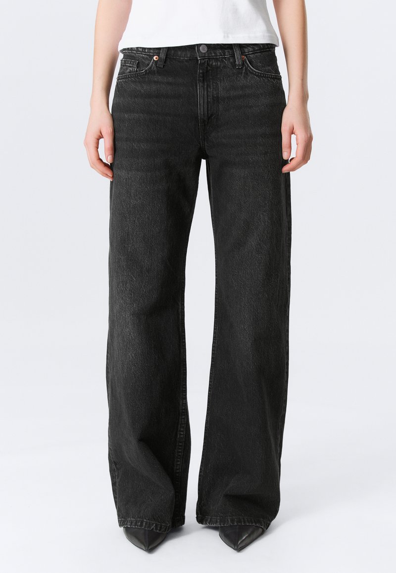 Γυναίκα που φοράει μαύρο ψηλόμεσο παντελόνι με φαρδιά πόδια, μαύρα μυτερά παπούτσια και λευκό τοπ, στέκεται μπροστά σε απλό φόντο.