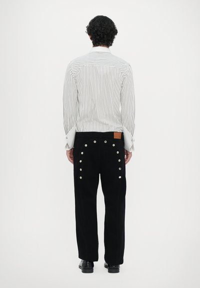 Camicia bianca a righe con ampi polsini, pantaloni neri con bottoni a pressione argento lungo i lati e una toppa in pelle marrone sulla vita.