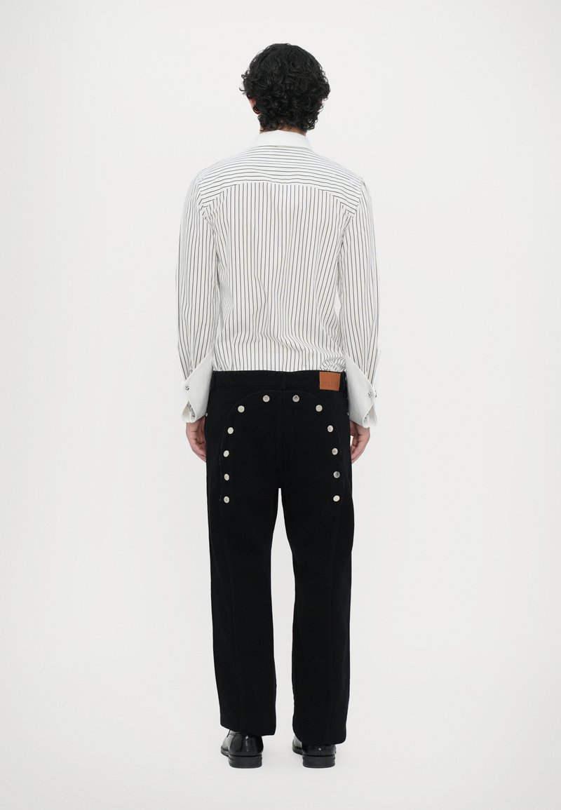 Chemise rayée blanche avec de larges poignets, pantalon noir avec des boutons-pression argentés sur les côtés, et une patch en cuir marron sur la taille.