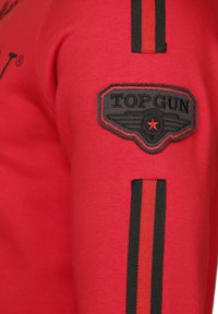 Röd sweatshirt med svarta accenter, med en broderad "TOP GUN"-patch och svarta randiga detaljer på ärmen. Mjuk tygtextur.