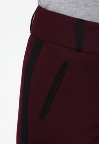 Pantalons bordeau avec une bande latérale noire et une poche zippée. Le tissu est lisse et extensible, avec une taille cintrée.