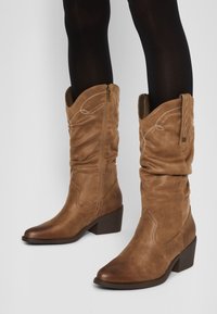 Botas vaqueras marrones de imitación cuero con un diseño arrugado, punta afilada y tacón apilado. Presentan costuras decorativas y cremallera lateral.