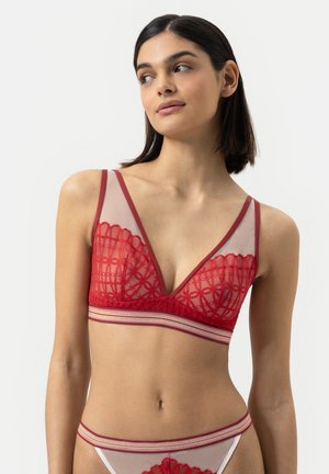 Jeune femme avec des cheveux noirs lisses portant un bralette en dentelle rouge et une culotte en tulle assortie, posant contre un fond gris clair.