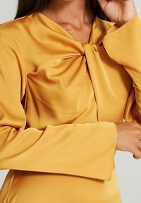 Gul satin topp med knutdetalj framtill, långa trumpetärmar och en slät textur. Figursydd passform med elegant drapering.