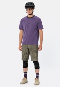 Chemise violette à manches courtes avec des accents noirs, associée à un short camouflage vert. Détails supplémentaires : genouillères et chaussettes violettes à motifs.