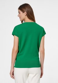 Grünes kurzärmliges Strickoberteil mit rundem Halsausschnitt und geripptem Saum, von hinten gezeigt, getragen mit hellen Hosen.