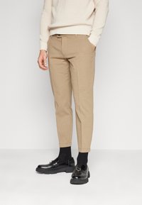 Chino beige con un design affusolato, realizzati in cotone testurizzato. Indossati con mocassini neri e calzini scuri, con i polsini arrotolati alla caviglia.