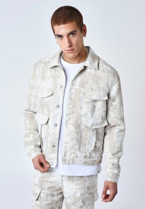 Veste en denim beige et blanche avec un motif texturé de taches de peinture. Comprend quatre poches avant et des boutons pression en argent.