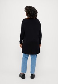Femme aux cheveux bouclés portant un long pull noir, un jean bleu clair et des chaussures noires cloutées, debout de dos sur un fond uni.