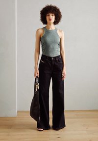 Zelený žebrovaný top na ramínka, kalhoty širokého střihu z tmavého denimu s vysokým pasem a černá ramenní taška se zlatým akcentem. Jednoduché pozadí a dřevěná podlaha.