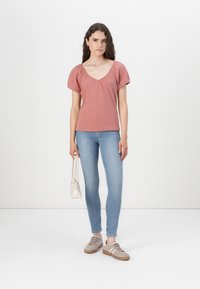 Bluză scurtă roz închis cu mânecă scurtă și decolteu în V, cu detalii plisate, purtată cu blugi skinny bleu deschis și adidași bej, ținând o geantă mică și ușoară.