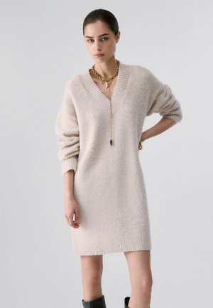Jeune femme portant une robe-pull beige oversize à col en V, un collier chaîne doré et des bottes noires, posant avec une main sur la hanche.