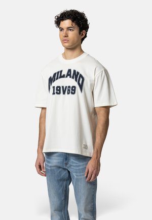Junger Mann mit lockigem Haar, der ein weißes "MILANO 19V69" T-Shirt und hellblaue Jeans trägt, steht vor einem einfachen weißen Hintergrund.