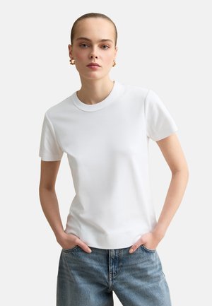 Mujer con el pelo recogido que lleva pendientes de aro dorados, una camiseta blanca lisa de manga corta y vaqueros azules con las manos en los bolsillos.