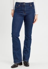 Dunkelblaue, ausgestellte Jeans aus Denim, mit fünf Taschen, gelben Nähten und einem vorderen Knopfverschluss. Getragen mit schwarzen Blockabsatzstiefeln.