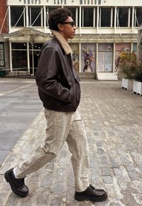 Rjava usnjena pilotska jakna z ovratnikom iz jagnječje volne, svetle teksturirane hlače in temni masivni čevlji, ki hodijo po tlakovanem kamnu.