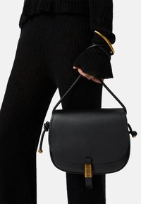 Borsa crossbody in pelle nera con forma arrotondata, dettagli dorati e una tracolla intrecciata. Abbigliamento in tessuto nero rigato parzialmente visibile.