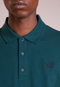Tyrkysové polo tričko se strukturovaným límcem, třemi knoflíky a kontrastním prošíváním. Obsahuje vyšívané logo na hrudi.