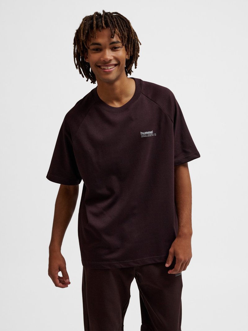 Junger Mann lächelt, trägt ein dunkelbraunes Hummel-T-Shirt und passende Hose, steht vor einem einfarbigen hellen Hintergrund.
