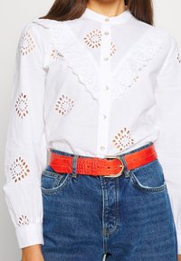 Blouse en coton blanc avec détails en broderie anglaise, col à volants et boutons en perles, associée à un denim bleu et une ceinture orange vif embossée crocodile.