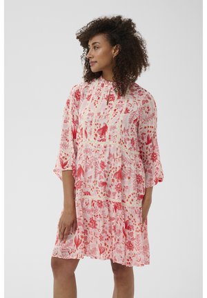 Femme aux cheveux bouclés portant une robe mi-longue à fleurs roses et rouges avec des manches trois-quarts, debout et regardant sur le côté.