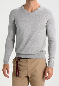 Pull gris avec un col en V et des poignets côtelés. Présente un petit logo sur la poitrine. Porté avec un pantalon beige clair et une ceinture rayée.