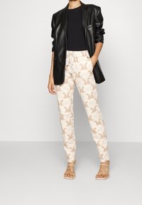 Schwarze Kunstleder-Blazer, schwarzes Top und cremefarbene Hose mit hellbraunem Tiermotiv. Klare Absatzsandalen runden das Outfit ab.