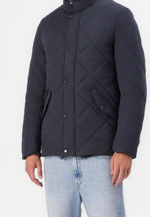 Man draagt een donkerblauw doorgestikt jack met drukknopen en klepzakken, gecombineerd met lichtblauwe spijkerbroek.