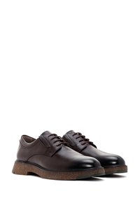 Chaussures en cuir texturé marron foncé à lacets avec des orteils ronds et de épaisses semelles couleur liège sur fond blanc.