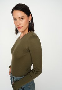 Maslinasto zelena rebrasta majica dugih rukava s okruglim izrezom i prilagođenim siluetom, uparena s svijetloplavim trapericama. Glatka tekstura, minimalistički dizajn.