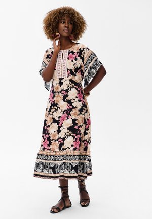 Kobieta z kręconymi włosami nosi czarną midi sukienkę w kwiaty z różowymi i beżowymi kwiatami, szerokie rękawy z wzorem oraz czarne sandały na sznurowadła.