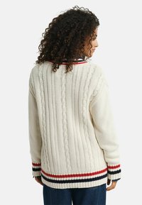 Frau mit lockigem Haar, die einen cremefarbenen, grob gestrickten Pullover mit rot, marineblau und weiß gestreiftem Rand trägt, von hinten gezeigt.