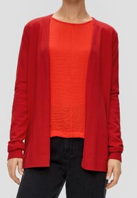 Cardigan rouge avec une texture douce, design devant ouvert et manches côtelées. Porté sur un haut rouge assorti et un jean noir.