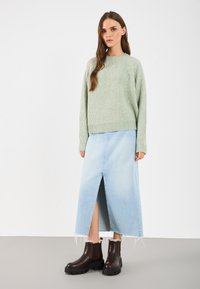 Une femme se tient debout, portant un pull en tricot vert clair, une jupe midi en denim bleu clair avec une fente à l'avant, et des bottes chunky marron foncé avec une doublure en fourrure.