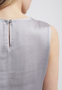 Haut sans manches gris soyeux avec détail d'encolure en forme de trou de serrure et fermeture par bouton au dos. Texture lisse avec une légère brillance.