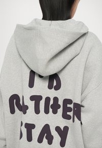 Grijze hoodie met grote, vette, donkerpaarse tekst op de rug die "IK BLIJF LIEVER" leest. Het materiaal lijkt zacht en met textuur.