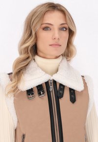 Beige neplederen vest met een witte, zachte kraag, zwarte riemen en gespen, en een ritssluiting aan de voorkant. Achtergrond is eenvoudig wit.