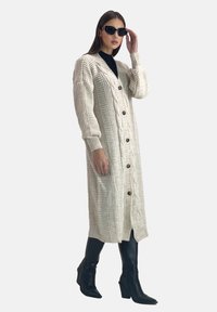 Lungo cardigan crema realizzato in maglia testurizzata, con motivo a treccia, polsini a coste e grandi bottoni, abbinato a stivali neri al ginocchio.