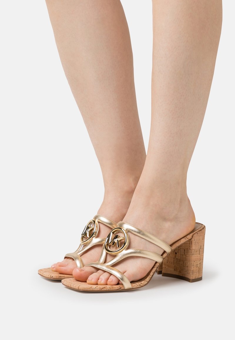 Pieds portant des sandales à talons hauts dorées avec talons blocs en liège et logo circulaire MK en métal sur les brides sur un fond uni.